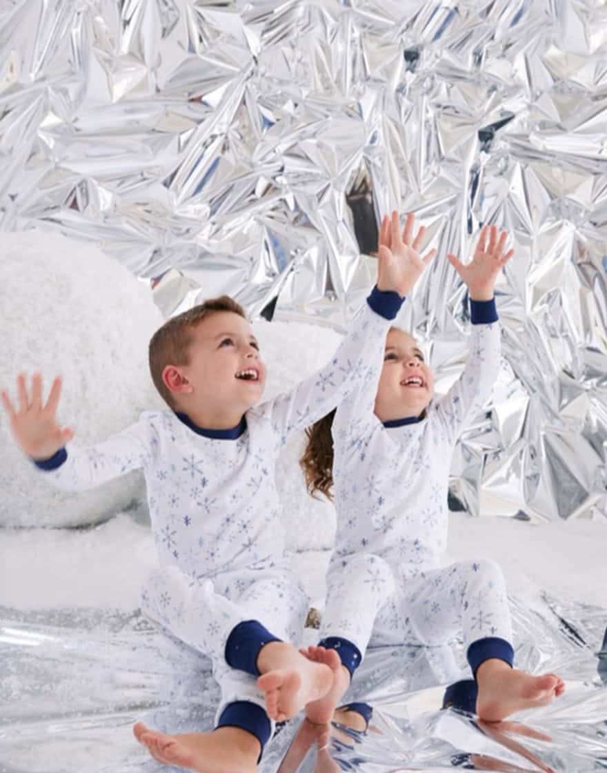 Two young children in printed pajamas playing in faux snow against backdrop made of silver mylar